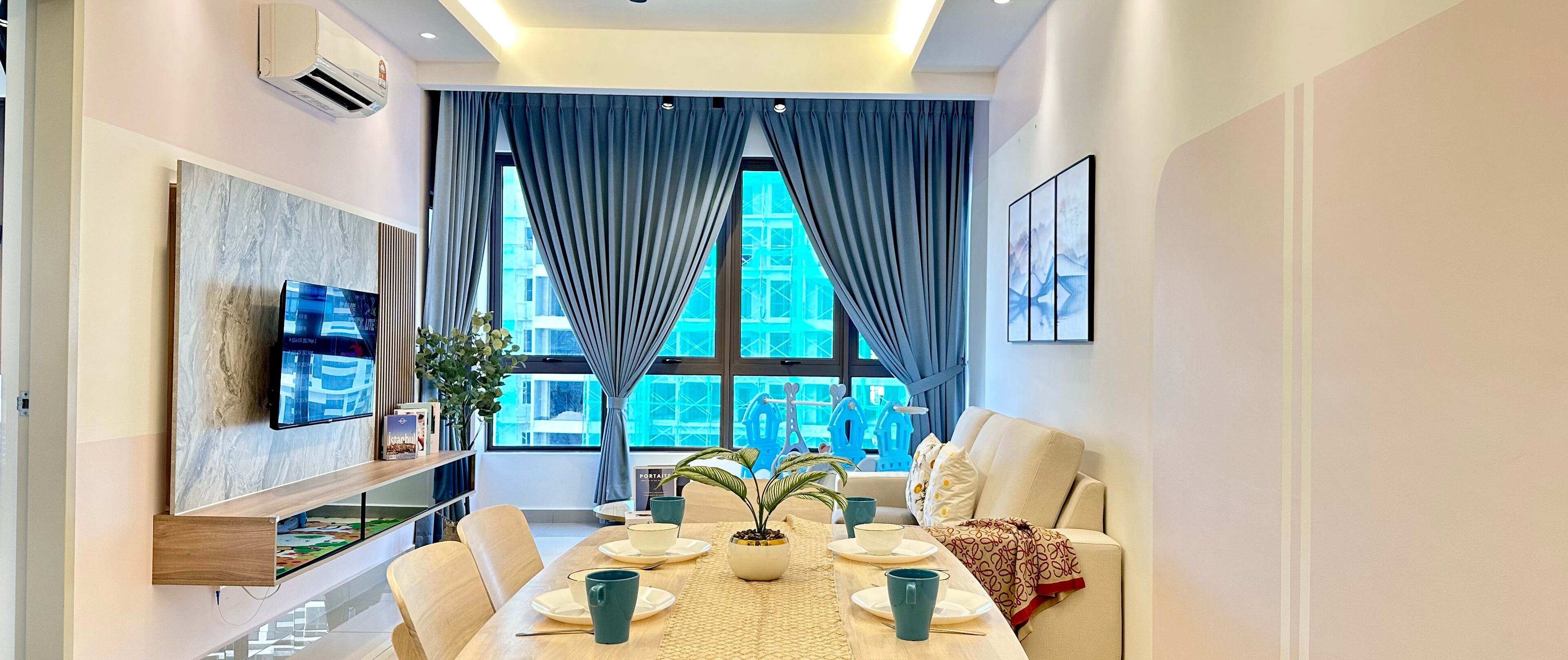 A modern living room with a dining area, large windows with curtains, a flat-screen TV, and a bookshelf.