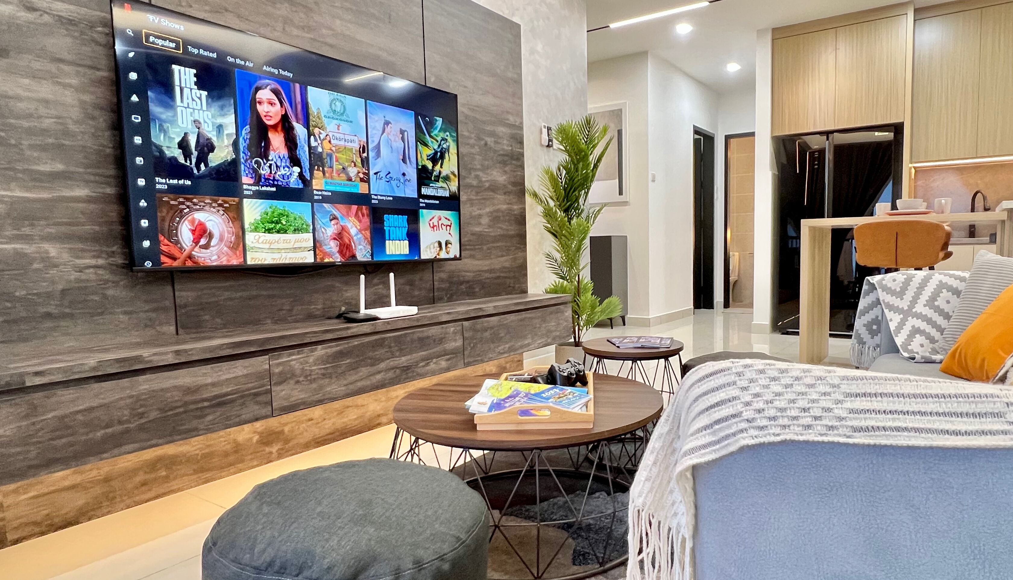 A modern living room with a flat-screen TV displaying various shows, a round coffee table with magazines, a grey sofa, and a wooden cabinet.