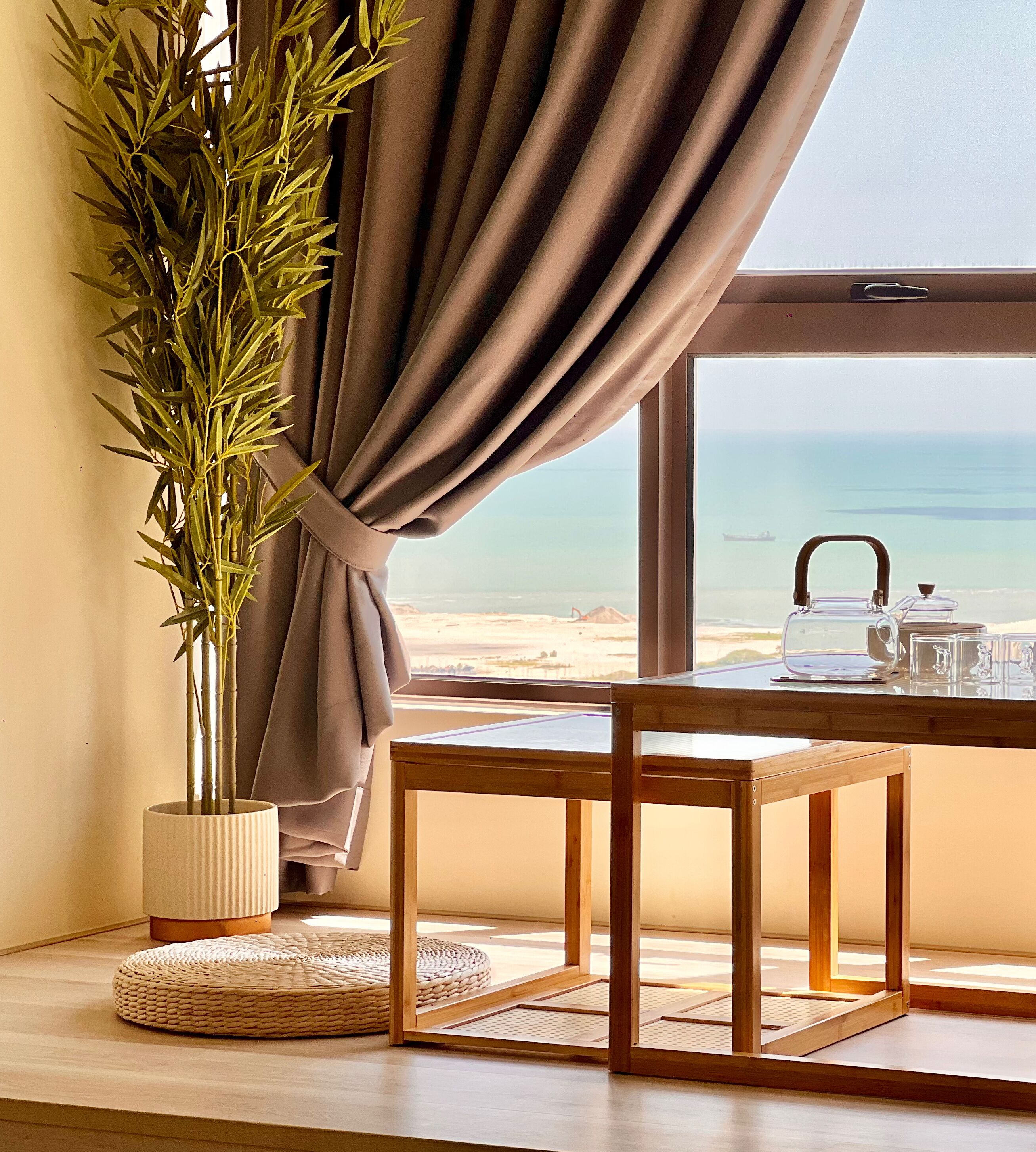 Een kamer met uitzicht op het strand, een tafel met theepot en kopjes, een bamboeplant en een geweven mat.