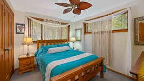 A bedroom with a wooden bed, a nightstand with a lamp, a ceiling fan, and a window with curtains.