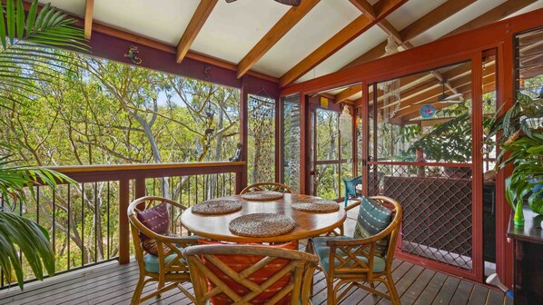 A wooden deck with a round table and chairs, surrounded by glass doors and windows offering a view of trees.