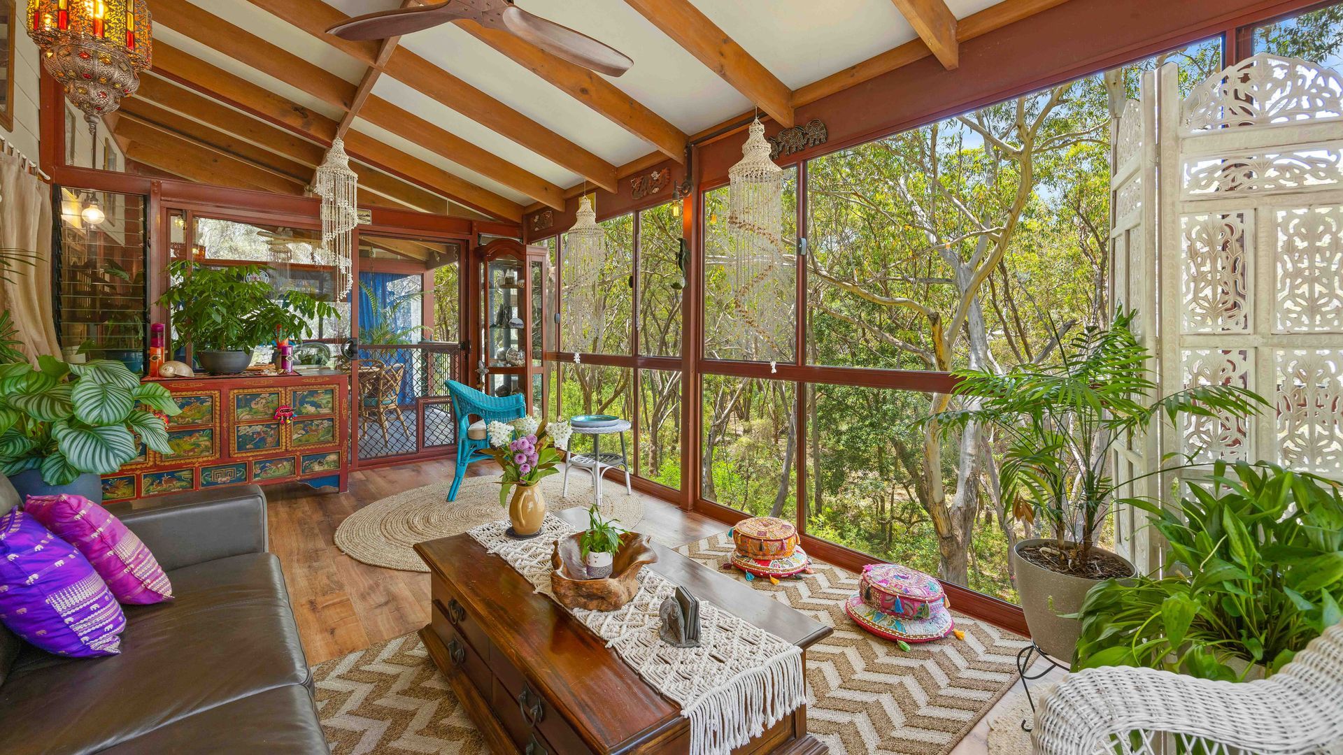 Amplia habitación con techo de madera, ventanas grandes y vista a árboles. Se observa un sofá, una mesa de comedor y varios objetos decorativos.