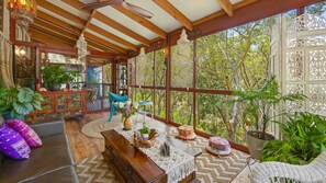 A spacious room with a wooden ceiling, large windows, and a view of trees. There is a sofa, a dining table, and various decorative items.