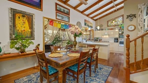 Un comedor con una mesa de madera, sillas y un jarrón de flores. Se observa una cocina al fondo con armarios y electrodomésticos.