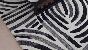 A neatly made bed with a green blanket and two pillows, a zebra-patterned rug, a bedside table with a lamp, and a chair.
