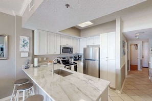 A modern kitchen with white cabinets, stainless steel appliances, and a marble island with bar stools. - Beachfront Views Indoor Pool Tidewater sleeps 6! (Orange Beach)
