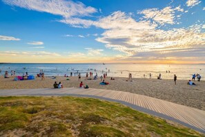 A beach with a wooden boardwalk leading to the water, people relaxing on the sand, and a sunset in the background.