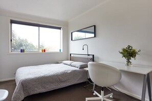 A bedroom with a bed, a desk, a chair, a window, and a mirror.