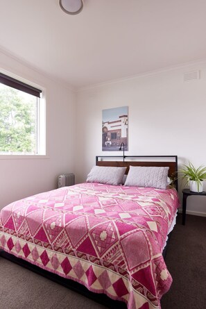 Un dormitorio con cama, ventana, radiador, un cuadro en la pared y una planta en maceta.