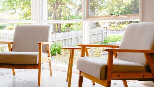 A room with two wooden chairs and a small round table, large windows with a view of a wooden fence and greenery outside.