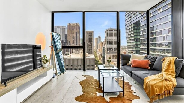 A modern living room with a large city view, a flat-screen TV, a glass coffee table, and a sofa with a throw blanket.
