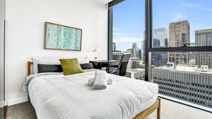 A modern hotel room with a large window offering a city view, a neatly made bed, a desk, and a framed artwork on the wall.