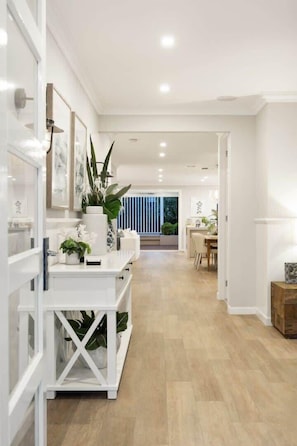 A modern, well-lit hallway with white cabinetry and a wooden floor, leading to a dining area with a view of greenery outside.