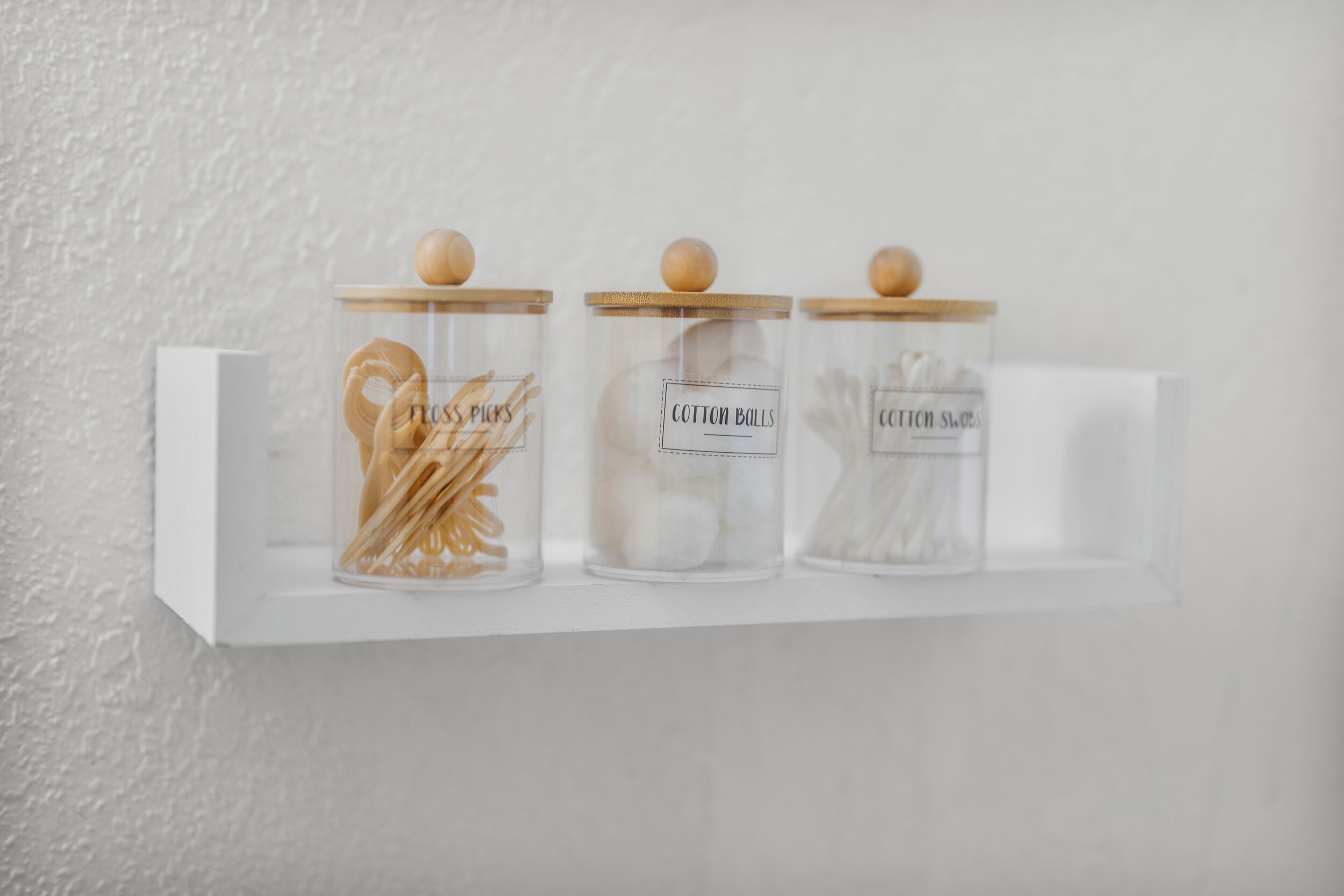 Tres frascos de vidrio con tapas de madera sobre un estante blanco frente a una pared con textura.