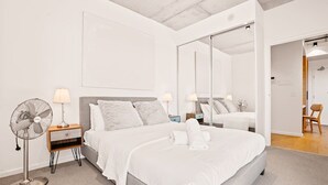 Une chambre d’hôtel moderne avec un grand lit, un ventilateur de chevet, une table de chevet, un miroir et une chaise en bois.