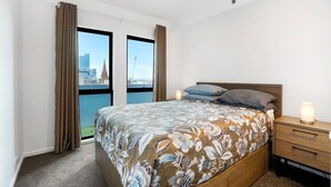 A hotel room with a large bed, two windows with curtains, a wooden nightstand, and a view of a cityscape.