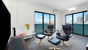 A modern office space with a view of the city skyline through large windows. There are three black leather chairs with cushions, a small round table, and a standing desk with a keyboard.