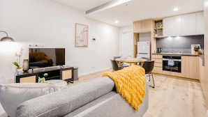 A modern living room with a sofa, TV, and dining area. A kitchen with wooden cabinets and appliances is visible in the background.