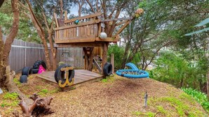 Una estructura de juego de madera con un tobogán y una plataforma, rodeada de árboles y mantillo.