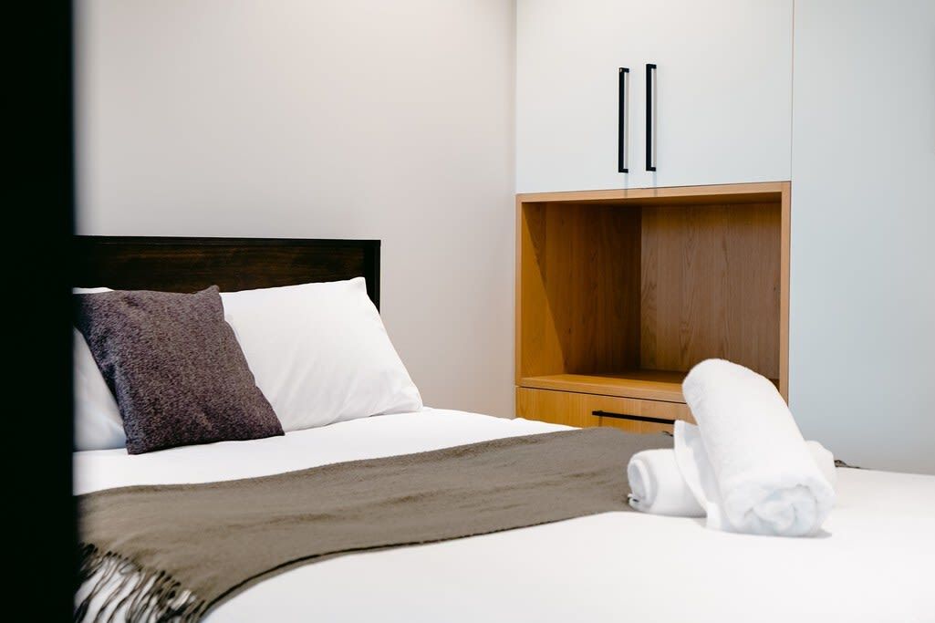 A hotel room with a bed, a wooden wardrobe, and a rolled-up towel on the bed.