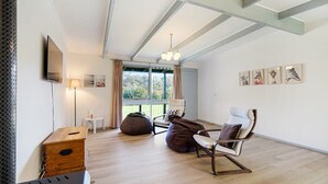 A modern living room with a wooden floor, a flat-screen TV mounted on the wall, a sofa, a chair, and a bean bag. There is a window with curtains, a lamp, and framed artwork on the wall. - Rockpool House Short stroll to  St. Andrews Beach (Saint Andrews Beach)