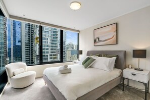 Una habitación de hotel moderna con una cama grande, una zona de estar, una mesita de noche y vistas al horizonte de la ciudad.