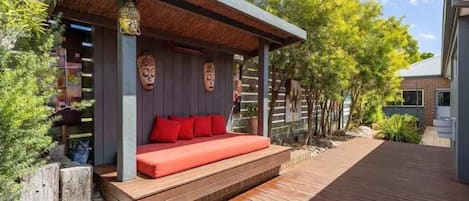 A covered outdoor seating area with a wooden deck, a built-in bench with red cushions, and decorative Buddha heads.