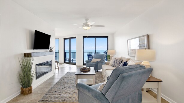 A spacious living room with a fireplace, a flat-screen TV, a ceiling fan, and a large window offering a view of the ocean. - 1BR Beachfront Beauty! (Orange Beach)