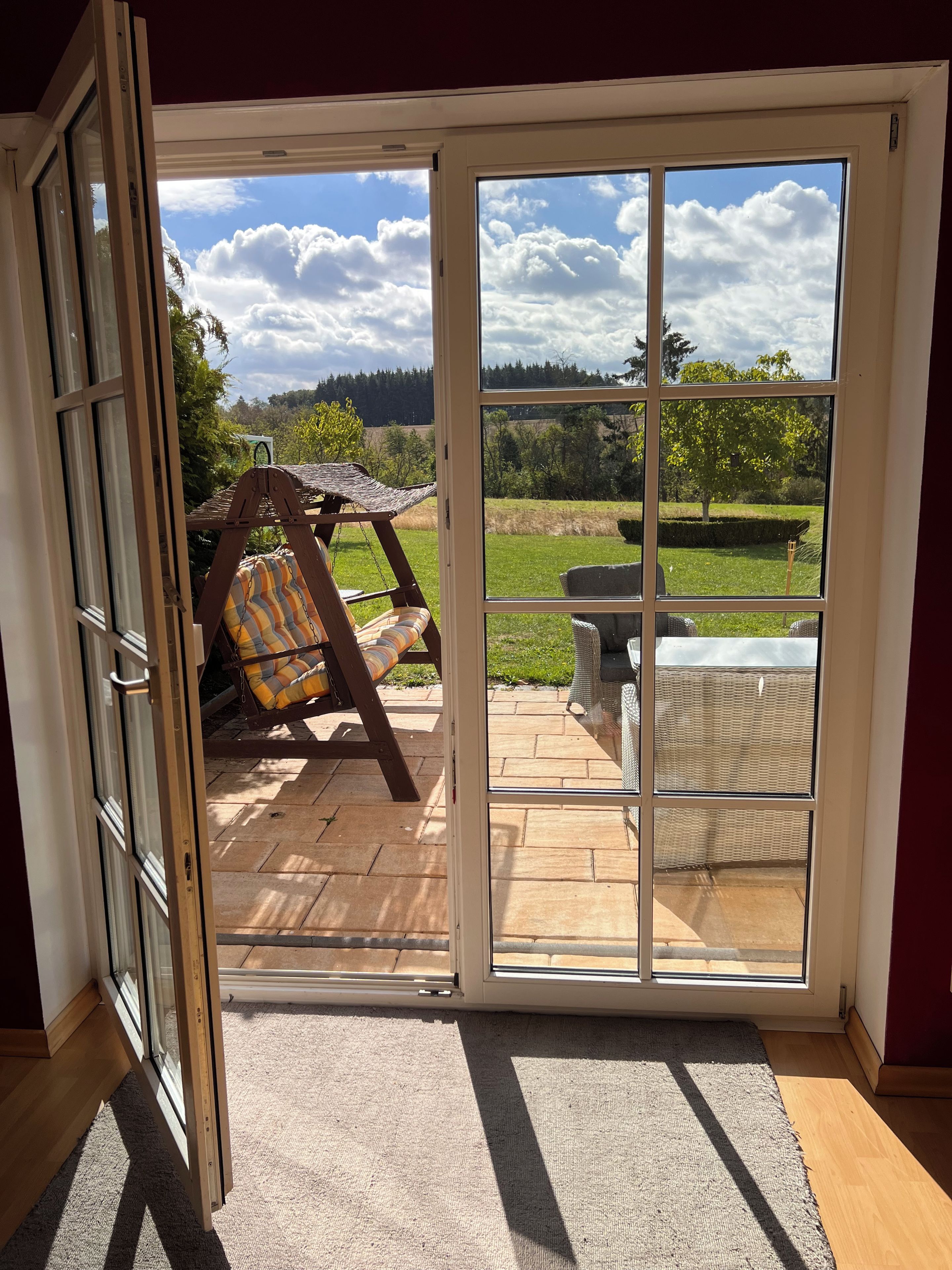A room with a sliding glass door leading to an outdoor patio with a swing chair, a wicker table, and a view of a green landscape.