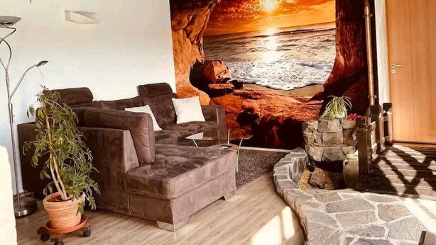 Una sala de estar con piso de piedra, un sofá, una planta en maceta y un mural de un atardecer sobre el océano.
