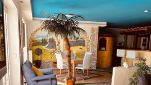 Amplia sala de estar con piso de piedra, una palmera en maceta grande y vista a un paisaje pintado a través de un arco.