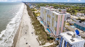 Un hotel frente a la playa con varios edificios altos, una piscina y una extensión de arena junto al océano.