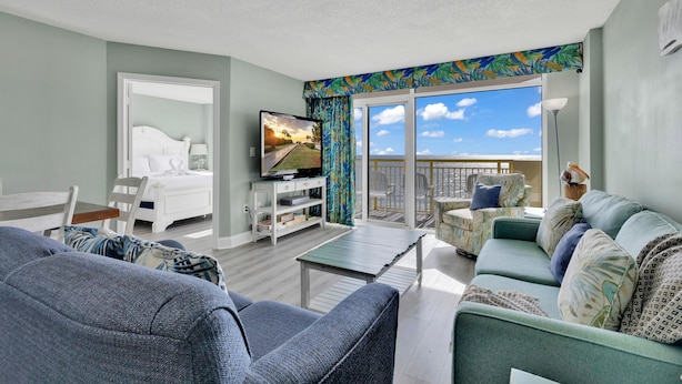 Habitación de hotel con balcón, cama, televisor y zona de estar con vistas al exterior.