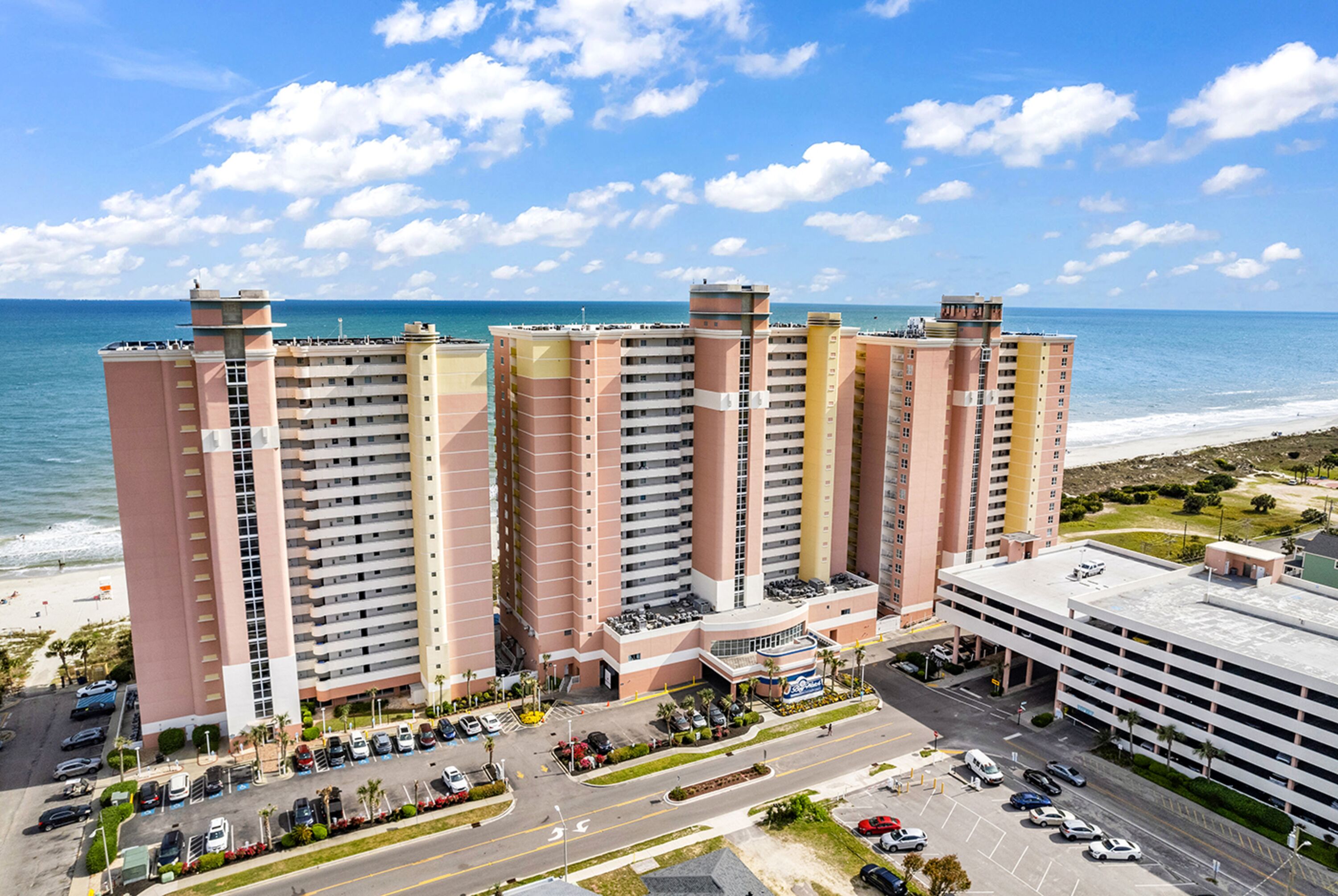 A high-rise hotel with multiple floors, a parking lot, and a view of the beach and ocean.