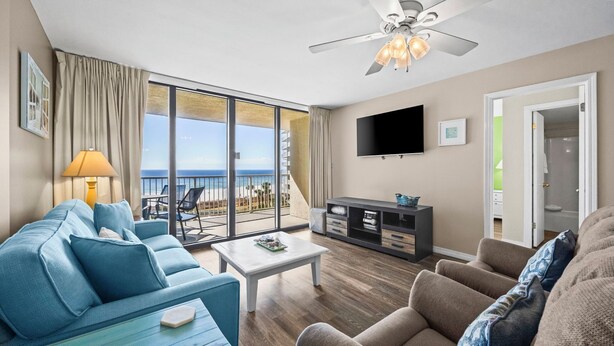 A spacious living room with a large window overlooking a balcony and the ocean. There is a ceiling fan, a flat-screen TV, and a seating area with a sofa and armchairs. - Gulf-front retreat with 3 pools, patio, and beach access! (Orange Beach)