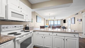 A modern kitchen with white cabinetry, a built-in oven, and a stove. There is a dining area with a view of the ocean. - Gulf-front retreat with 3 pools, patio, and beach access! (Orange Beach)