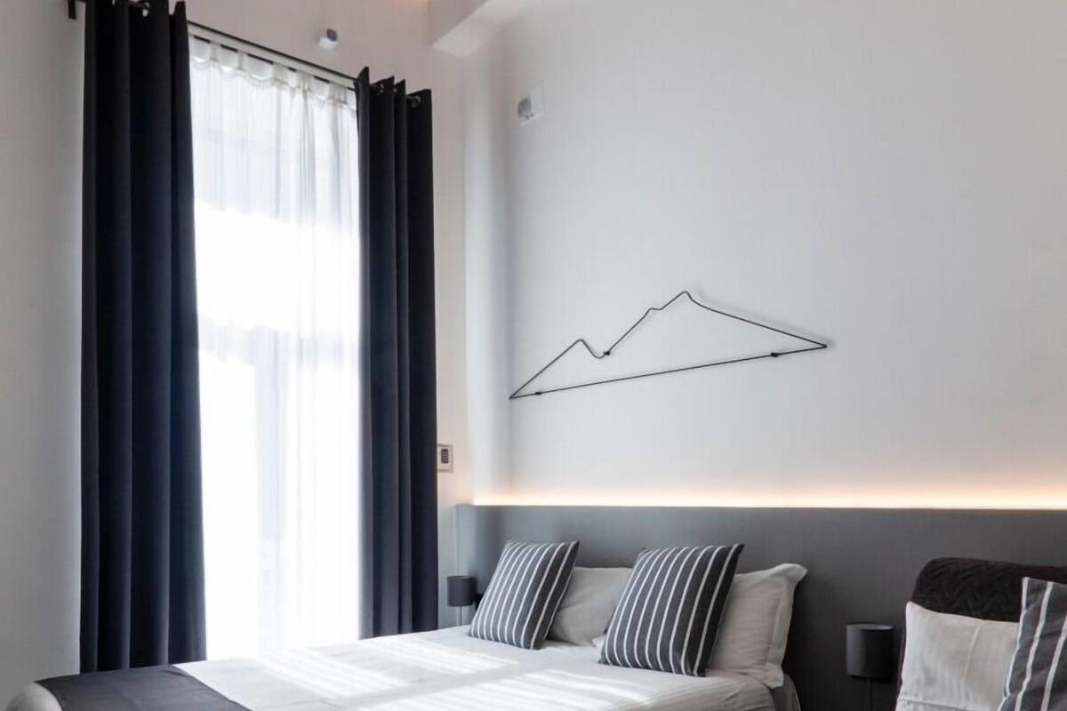 A modern hotel room with a bed, a sofa, a wall-mounted light, and a ceiling with exposed beams.