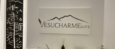 Un espace d’accueil avec une enseigne fixée au mur indiquant « Vesucharm Esuite », un bureau avec une lampe, un vase contenant des tiges de roseau et un récipient en verre avec une cuillère.