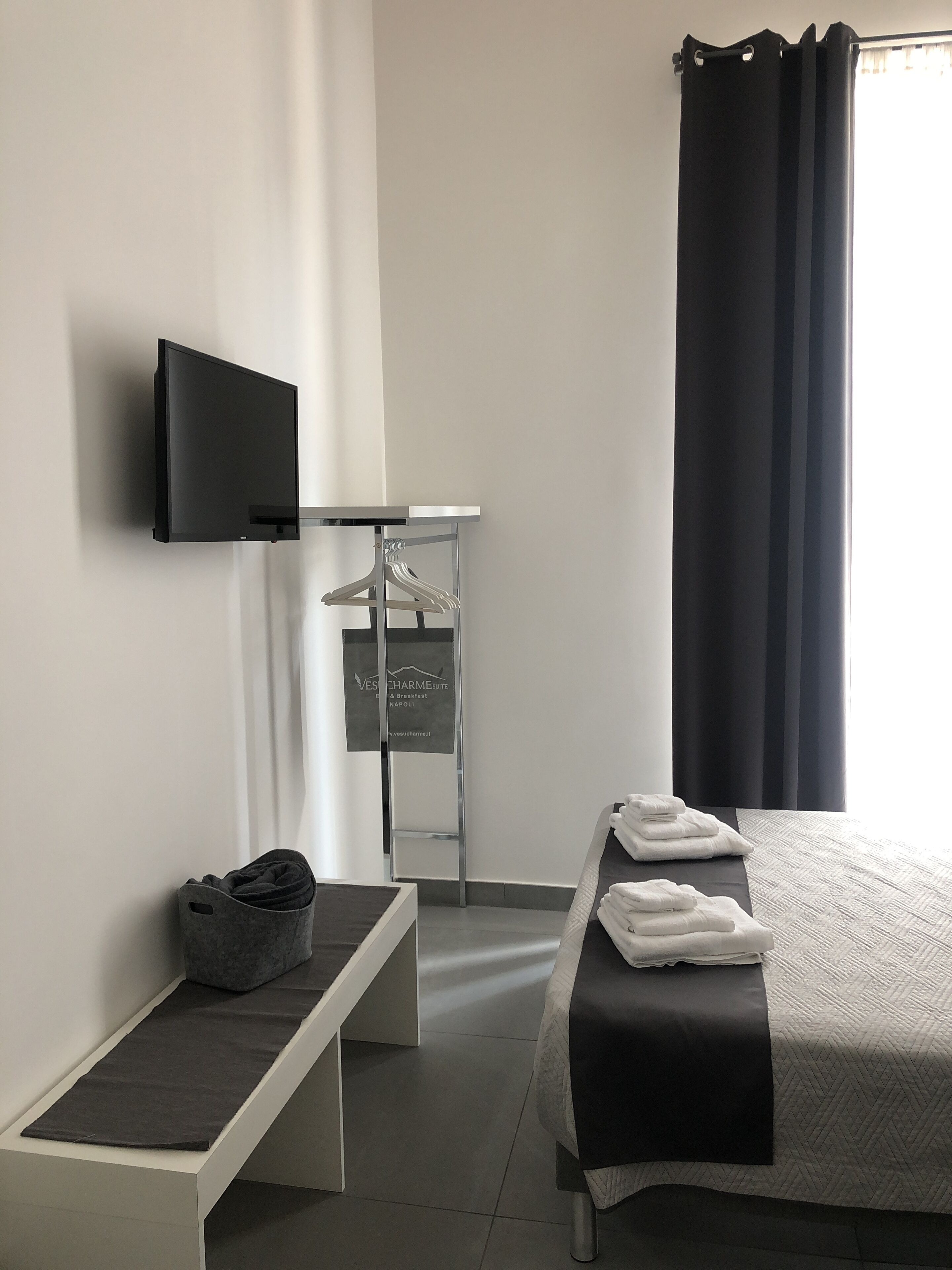 Quarto de hotel moderno com uma cama, televisão fixada na parede, um banco, um cabideiro e um aviso publicitário do hotel.