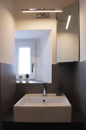 A modern bathroom with a rectangular sink, a mirror above it, and a window providing natural light. - VESUCHARME Suite Luxury Room (Naples)