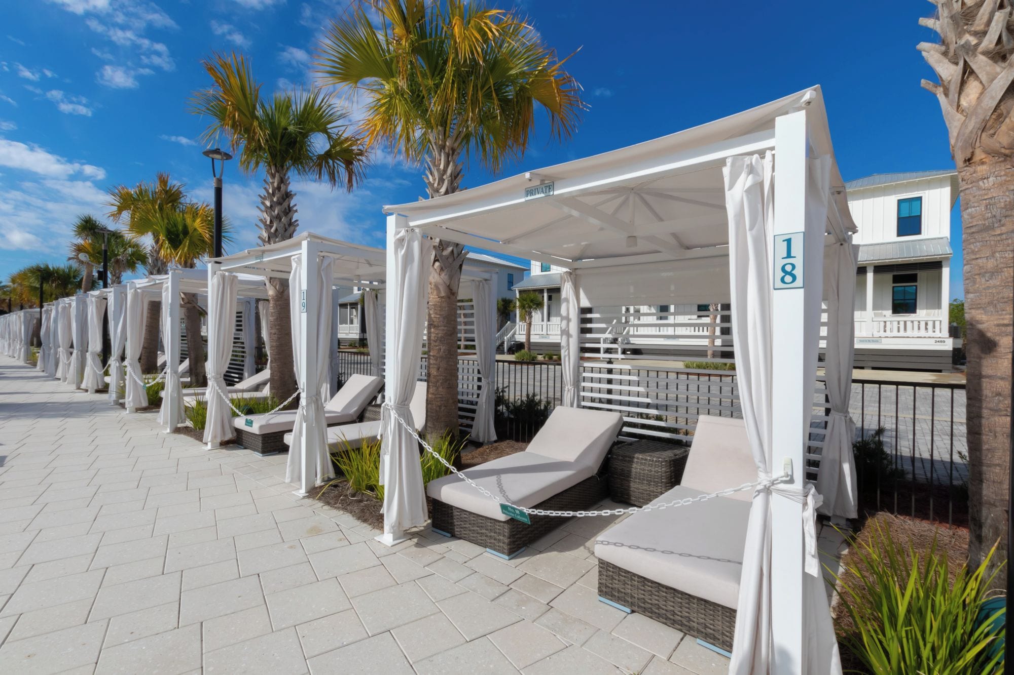 A row of private cabanas with white canopies, numbered 18, along a paved walkway with palm trees in the background.
