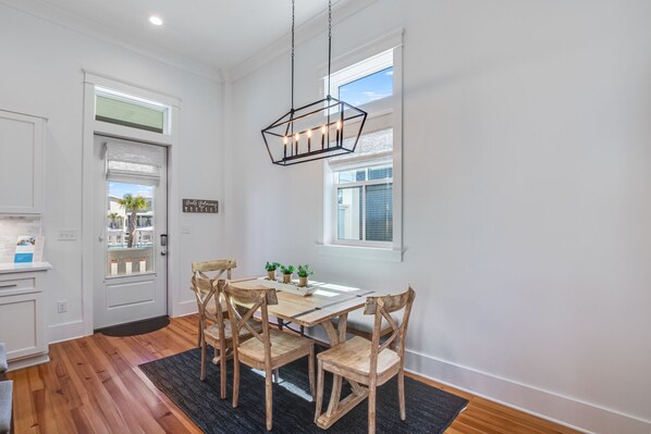 Un comedor con mesa y sillas de madera, una lámpara colgante y vista al exterior a través de una ventana.