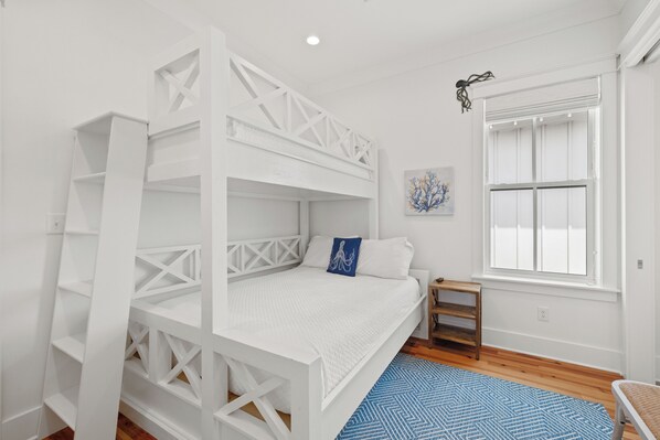 A white bunk bed with a blue pillow, a small wooden side table, a blue patterned rug, and a window with white shutters.