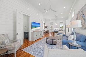 A spacious living room with a large TV, a dining area, and a kitchen in the background.
