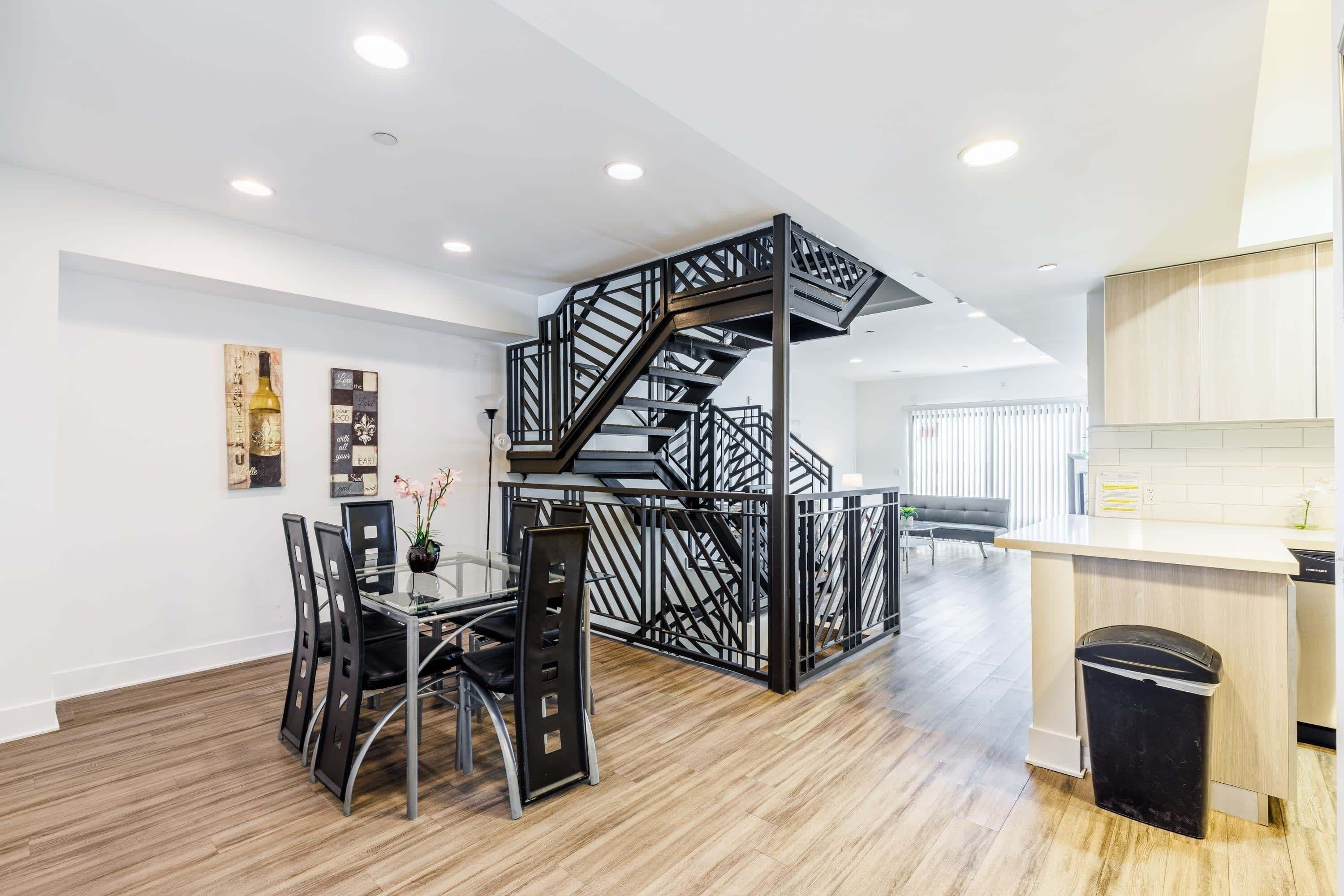 A modern kitchen with a wooden floor, a black staircase, and a dining area with a glass table and chairs.