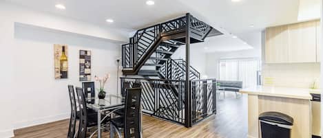 A modern kitchen with a wooden floor, a black staircase, and a dining area with a glass table and chairs.