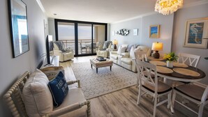 A spacious living room with a balcony, a dining area with a round table, and a large chandelier. - Ocean Creek Kk-14 2 Bedroom Condo by RedAwning (Myrtle Beach)