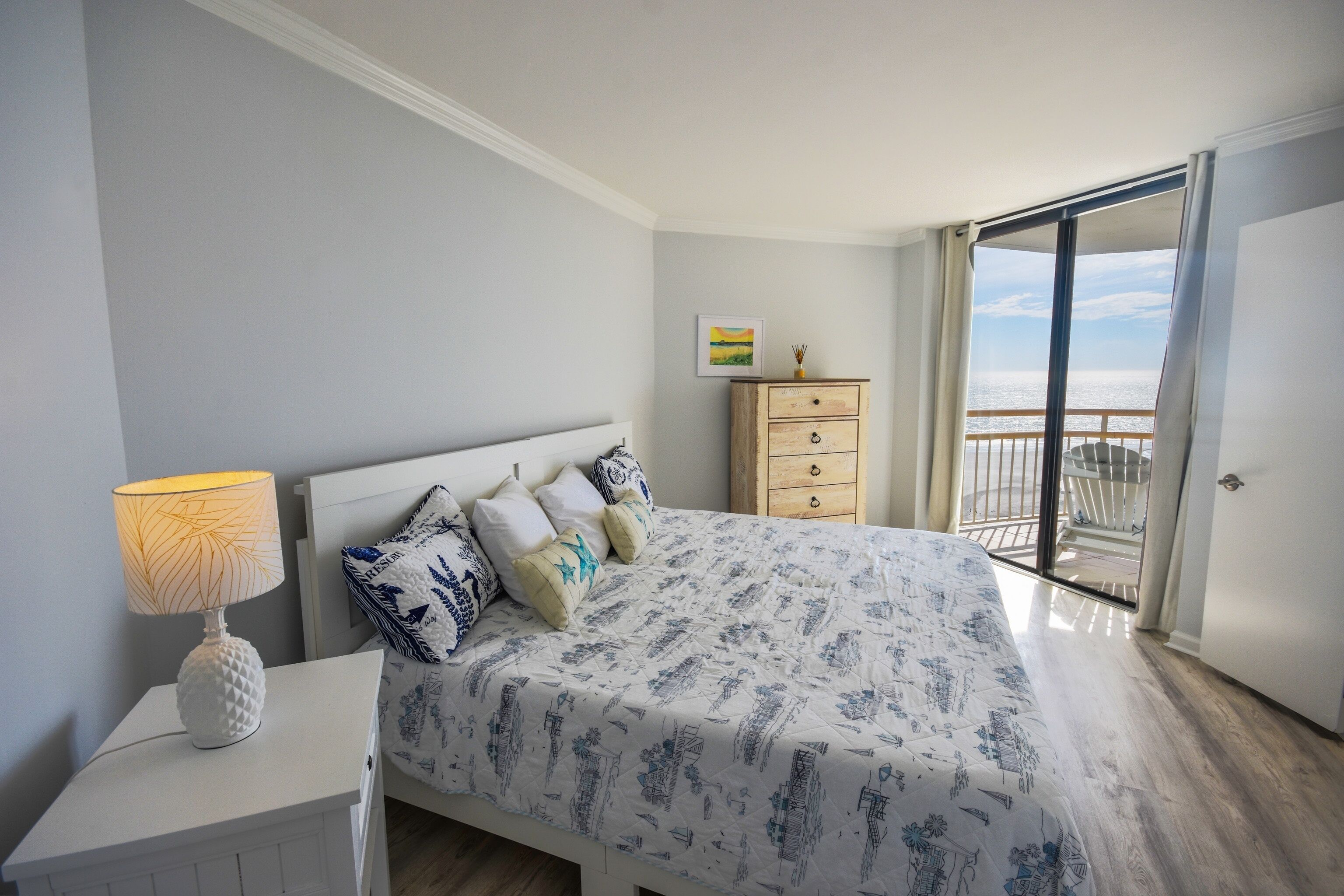 Ein Schlafzimmer mit Bett, Schminktisch, Nachttisch mit Lampe, einem Wandbild und einem Balkon mit Blick.