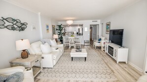 A modern living room with a white sofa, a coffee table, and a TV stand. There is a kitchen in the background with white cabinets and a chandelier. - Ocean Creek Kk-14 2 Bedroom Condo by RedAwning (Myrtle Beach)