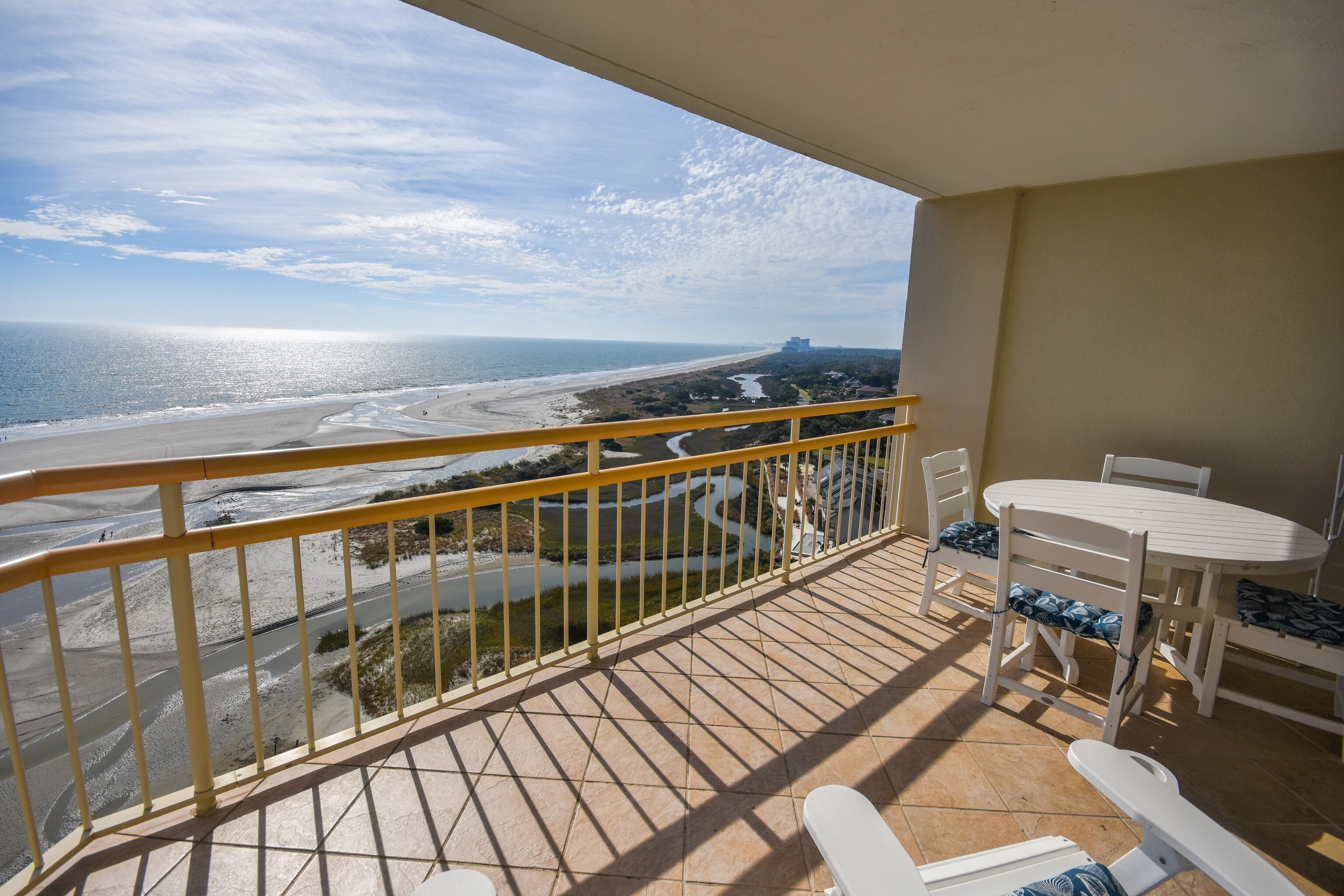 A balcony with a view of a beach and river. There is a table and chairs on the balcony.